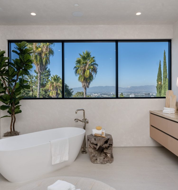 Modern bathroom with a freestanding tub, large window with a view of palm trees and mountains, and minimalist decor.